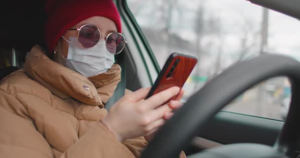Een jonge vrouw in een auto met een medisch masker op haar gezicht schrijft een sms op haar mobiele telefoon. Het uitzicht en het verspreidingsgebied van het virus. Pandemie, quarantaine, coronavirus, isolatie — Stockvideo