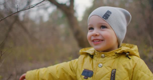 Feliz niño risueño y sonriente de 2 años en el bosque mira a la cámara. Encantador chico lindo con pestañas grandes. Un niño con una chaqueta y un sombrero amarillos — Vídeos de Stock