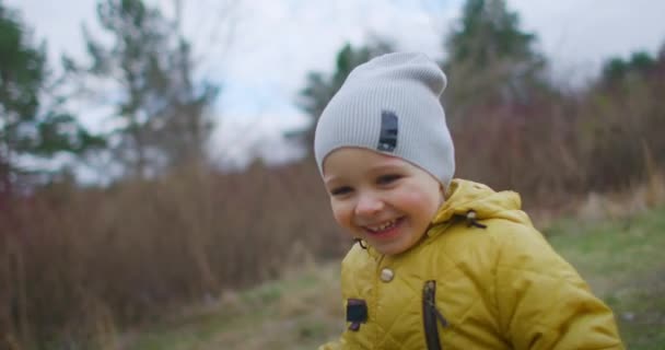 Happy Child Running In The Woods. Um rapazinho feliz a brincar no parque no Outono. Movimento lento: O menino com o casaco amarelo olha para a câmera Risos e sorrisos. Feliz infância livre . — Vídeo de Stock