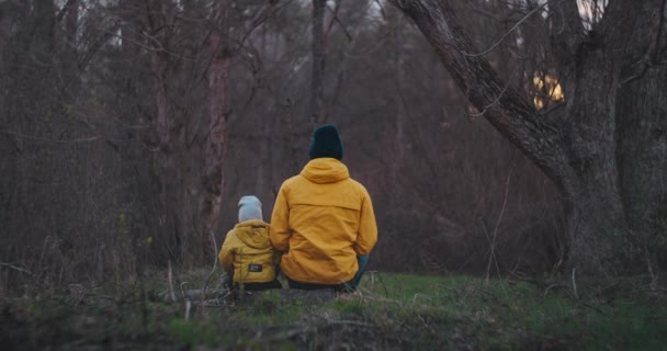 Zeitlupe: Rückansicht Vater erzählt etw für Sohn, während er auf einem Baumstamm sitzt. Glücklicher Vater und Sohn im Gespräch im Park. Vater und Sohn in gelben Jacken in der Natur-Herbstlandschaft. — Stockvideo