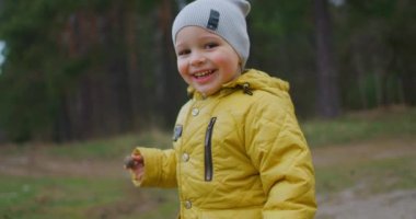 Ormanda koşan mutlu çocuk. Mutlu küçük çocuk sonbaharda Park 'ta oynuyor. Ağır çekim: Sarı Ceketli Çocuk kameraya bakıp gülüyor ve gülümsüyor. Mutlu özgür çocukluk.