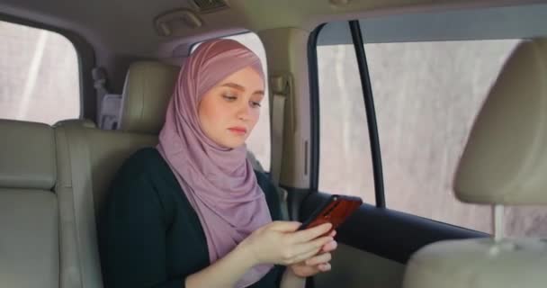 Oung krásná muslimka v hidžábu v autě spolujezdce zadní sedadla psaní na mobilní telefon s vážným výrazem. Internetová banka. Businesswoman pracuje na smartphone na zadním sedadle crowdsourced taxi — Stock video