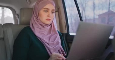 Hijab 'daki iş kadını arabanın arka koltuğunda dizüstü bilgisayar kullanıyor. Modern Arap iş kadını dizüstü bilgisayar ve taksi kullanıyor. Takside oturan ve ofise giden meşgul bir kadın.