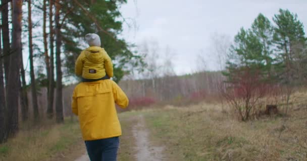 Zeitlupe: Vater legte Jungen auf seine Schultern und trug ihn durch Vorstadtpark. Der Vater trägt seinen Sohn auf Schultern und geht im Park spazieren. Sohn und Vater in gelben Jacken spazieren im Wald. — Stockvideo