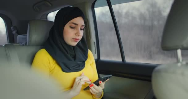 Oung bela mulher muçulmana no hijab no assento traseiro do passageiro do carro digitando no telefone celular com expressão séria. Banco da Internet. Empresária trabalhando no smartphone no banco de trás do táxi crowdsourced — Vídeo de Stock