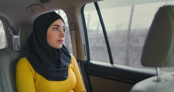 히잡에 앉아 있는 젊은 근육질 여성 이 차 뒷좌석에 앉아 있습니다. 이슬람 여성의 날, 뒷자리에서 꿈꾸고 있습니다. 히잡을 입은 젊은 여자가 창밖을 내다본다. — 비디오