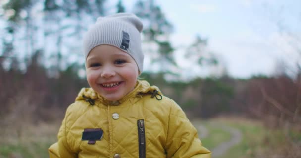 Joyeux enfant qui court dans les bois. Joyeux petit garçon jouant dans Park en automne. Ralenti : The Boy in the Yellow Jacket Runs regarde la caméra Laughs and Smiles. Heureuse enfance libre .
