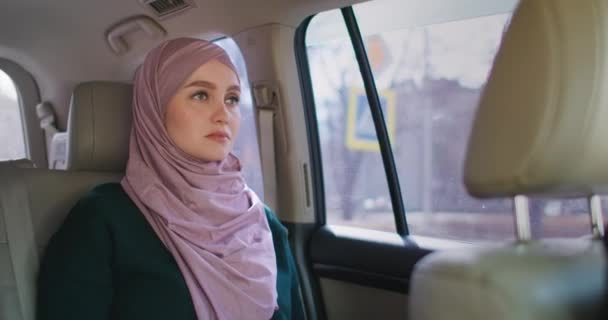 Seorang wanita Arab yang serius dan bijaksana di dalam mobil di kursi penumpang. Naik ke mobil dan melihat ke luar jendela. Seorang wanita Muslim dalam naik jilbab di bagian belakang mobil — Stok Video