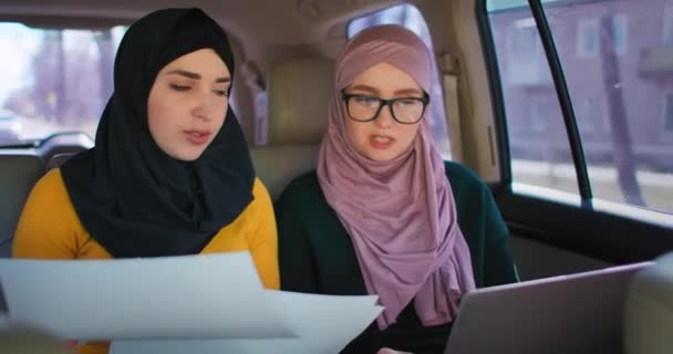 Jovens empresários árabes com dois laptops nos assentos traseiros do carro. Trabalho em equipe em movimento 2 Mulher de negócios muçulmana moderna em automóvel. Dois colegas sauditas da empresária que trabalham com o portátil no carro — Vídeo de Stock