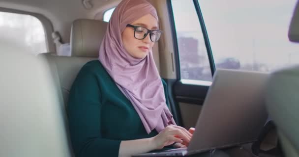 Mujer de negocios en Hijab utiliza portátil sentado en el asiento trasero del coche. Moderna mujer de negocios árabe usando computadora portátil y taxi a caballo, mujer ocupada sentada en taxi e ir a la oficina — Vídeos de Stock