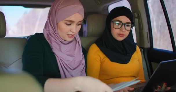 Jóvenes empresarias árabes con dos portátiles en los asientos traseros del coche. Trabajo en equipo en movimiento 2 Moderna mujer de negocios musulmana en el automóvil. Dos compañeras de negocios sauditas trabajando con el ordenador portátil en el coche — Vídeos de Stock