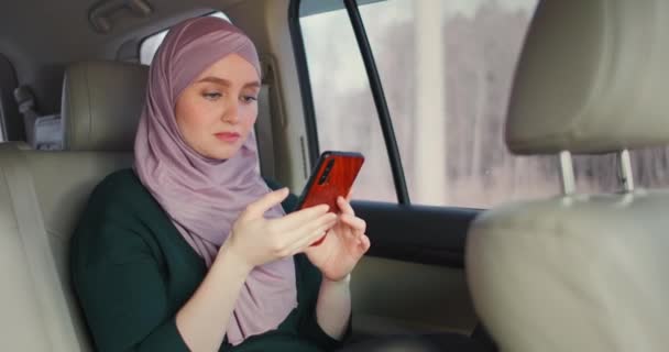 Oung bela mulher muçulmana no hijab no assento traseiro do passageiro do carro digitando no telefone celular com expressão séria. Banco da Internet. Empresária trabalhando no smartphone no banco de trás do táxi crowdsourced — Vídeo de Stock