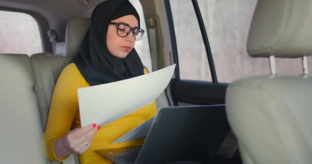 Business Arabian Woman Looking Financial Papers in vehicle (en inglés). Mujer en hiyab leyendo documentos en un coche. Usa un portátil y revisa los documentos. Mujer musulmana moderna de negocios . — Vídeos de Stock