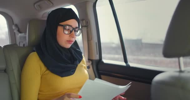 아랍 여성, 금융 신문을 자동차로 보고 있다. 히잡에 있는 여자가 차 안에서 문서를 읽고 있어요. 노트북을 사용하여 문서를 확인하 십시오. 현대 회교 사업 여성. — 비디오