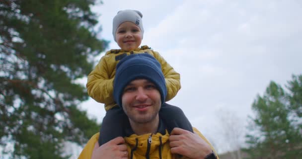 Zeitlupe: Vater legte Jungen auf seine Schultern und trug ihn durch Vorstadtpark. Der Vater trägt seinen Sohn auf Schultern und geht im Park spazieren. Sohn und Vater in gelben Jacken spazieren im Wald. — Stockvideo