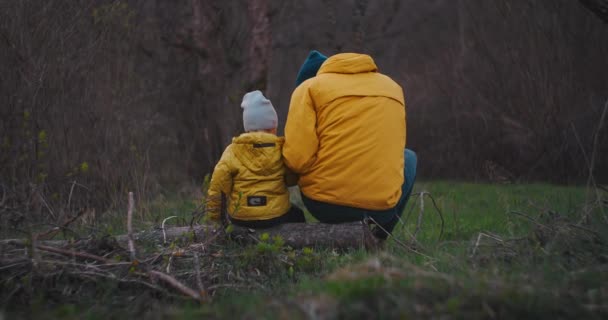 Zeitlupe: Rückansicht Vater erzählt etw für Sohn, während er auf einem Baumstamm sitzt. Glücklicher Vater und Sohn im Gespräch im Park. Vater und Sohn in gelben Jacken in der Natur-Herbstlandschaft. — Stockvideo