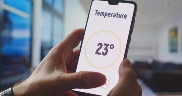 Ελέγχοντας τη θερμοκρασία του σπιτιού χρησιμοποιώντας ένα έξυπνο σπίτι ελέγχου χρησιμοποιώντας το κινητό τηλέφωνο. Hand κατέχει ένα smartphone με εφαρμογή στο σαλόνι για τον έλεγχο των συστημάτων ασφαλείας — Αρχείο Βίντεο