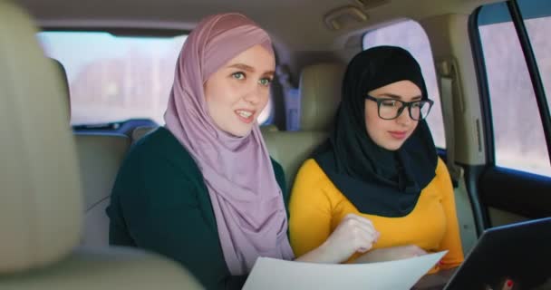 Dos mujeres de negocios viajan en un coche y discuten un plan de desarrollo de negocios. Mujeres islámicas en hiyabs miran documentos — Vídeos de Stock