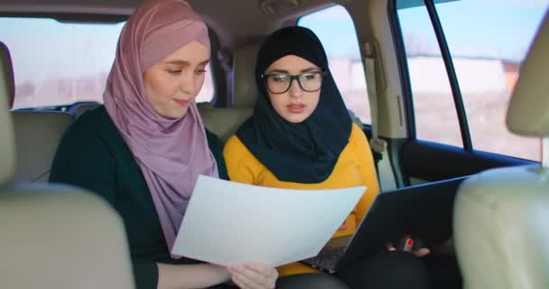 Twee zakenvrouwen rijden in een auto en bespreken een business development plan. Islamitische vrouwen in hijabs kijken naar documenten — Stockvideo
