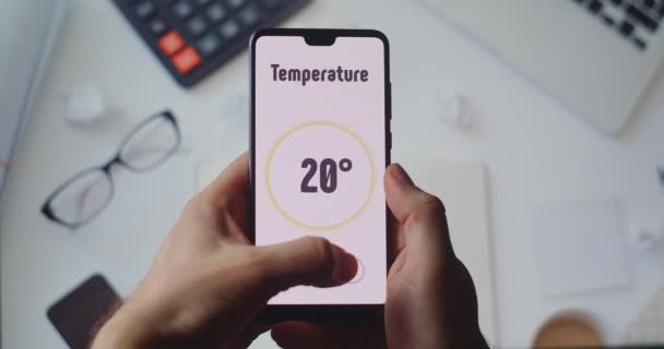 Gestisci le tecnologie smart home dall'ufficio. Sullo sfondo del desktop, le mani tengono uno smartphone con l'app Smart Home e impostano la temperatura e l'allarme in casa — Video Stock