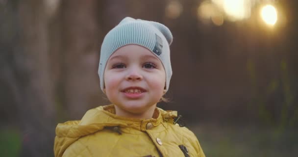 Ralenti : Jeune garçon vêtu d'une veste jaune Explorez le monde qui l'entoure assis dans la forêt au soleil. Profitez et riez dans les bois. Enfance joyeuse gratuite dans le parc dans la nature — Video