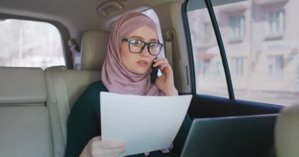 Una donna d'affari musulmana cavalca in macchina e guarda i documenti e parla al telefono. Condurre affari a distanza dalla vostra auto. Donna d'affari . — Video Stock