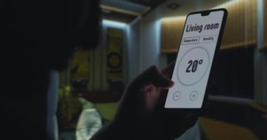 Akıllı bir ev kontrolü kullanarak evin sıcaklığını kontrol etmek için cep telefonu kullanmak. Oturma odasında güvenlik sistemlerini kontrol eden bir uygulama olan akıllı telefonu tutan el.