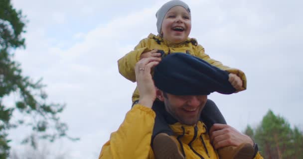 Hijo y padre son traviesos juntos. El niño se sienta en el cuello de sus padres y se quita el sombrero. Feliz risa y alegría del niño lepra. Diversión familiar juntos. Padre, hijo diviértete en el parque — Vídeos de Stock