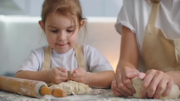 エプロンのキッチンで自宅の娘と母親は、圧延ピンで小麦粉をロールアウトし、一緒に手で生地を彫刻を注ぐ。子供と一緒に時間を過ごし、経験を積む. — ストック動画
