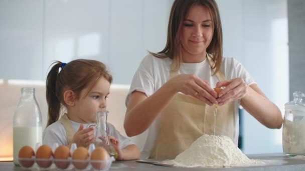 Önlüklü bir anne ve kız bir yumurta kırıyor ve hamur yapmak için unun içine atıyorlar. — Stok video