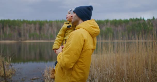 De vader houdt zijn zoon in zijn armen en kijkt samen naar het meer. Familie nabijheid van vader en kind. Vaderschap en levensstijl in een authentieke omgeving van natuur. — Stockvideo