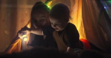 Küçük sevimli çocuğun masal kitabındaki bir şeye işaret ettiği ve el feneri kullanarak birlikte okurken ilgili anneyle konuştuğu yakın plan. Çocuklar için Noel süslemeleri, ışıklar ve çelenkler.