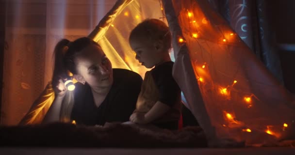 Крупним планом знімок милий маленький хлопчик вказує на щось в казковій книзі і розмовляє з дбайливою матір'ю, читаючи разом за допомогою ліхтарика. Різдвяні прикраси для дітей, вогні та гірлянди . — стокове відео