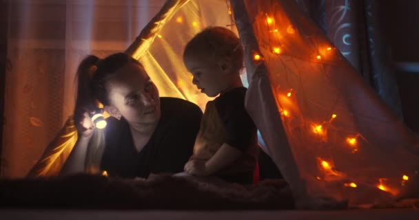 Primer plano de lindo niño apuntando a algo en el libro de cuentos de hadas y hablando con la madre cariñosa al leer juntos utilizando la linterna. Decoraciones navideñas para niños, luces y guirnaldas . — Vídeo de stock