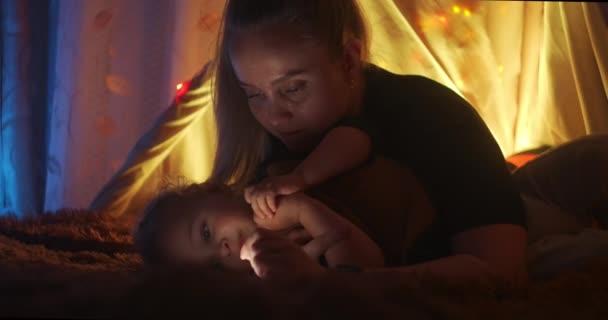 Parenthood, mom holds a mobile phone with a flashlight and reads book for sons in the wigwam. Little boy playing in children tent at home. Mother reading fairy tale book — Stock Video