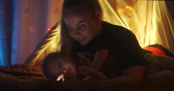 Close up shot of cute little boy pointing at something in fairy tale book and talking to caring mother when reading together using flashlight. Decorações de Natal para crianças, luzes e guirlandas . — Vídeo de Stock