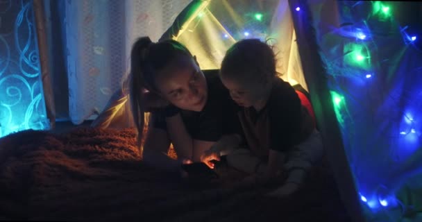 La madre legge un libro al figlio la sera in una tenda giocattolo in salotto. Mamma leggendo con ragazzo in cena in camera da letto a casa. Famiglia alla vigilia di Natale . — Video Stock