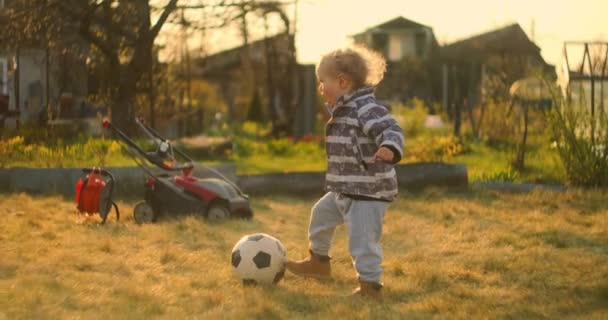 Ένα μικρό αγόρι κλωτσάει μια μπάλα ποδοσφαίρου με το πόδι του στο γήπεδο πίσω από το σπίτι. Ένας μικρός ποδοσφαιριστής σε αργή κίνηση. Πίσω αυλή. — Αρχείο Βίντεο