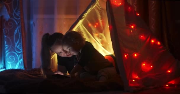 La favola della buonanotte. Mamma e figlioletto felici leggono un libro in tenda a casa. Leggere un libro sotto una coperta con una torcia in una stanza buia di notte. donna caucasica godendo di tempo in famiglia con suo figlio . — Video Stock