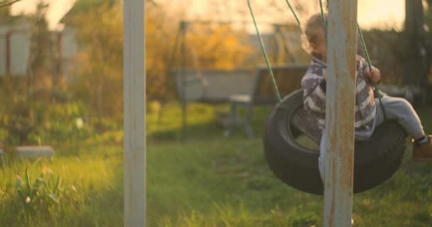 Un niño se balancea en un columpio al sol en el patio trasero. Feliz bebé. Movimiento lento — Vídeos de Stock