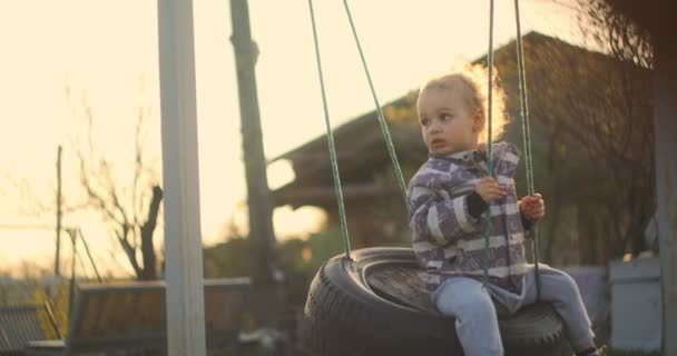 Un petit garçon se balance sur une balançoire au soleil dans le jardin. Joyeux bébé. Mouvement lent — Video