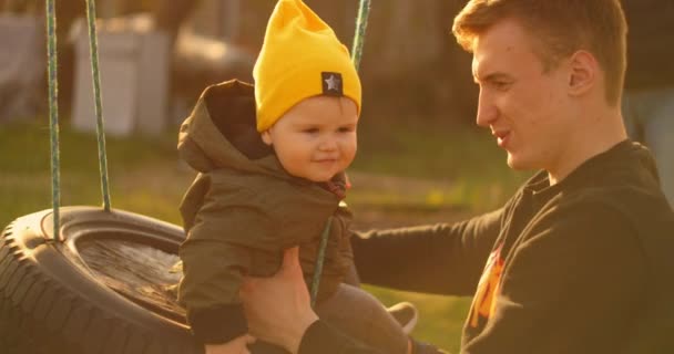 Papà felice fa rotolare un figlio allegro su un'altalena in un parco su una quercia. Il concetto di famiglia felice. Padre single. rallentatore padre e figlio su un swing il giorno del padre . — Video Stock