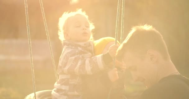 スローモーション:幸せな若い父親がスイングに彼の息子を押しています。父と2人の息子が庭で遊んでいると、美しい太陽の光で木からぶら下がってタイヤスイング。2人の兄弟と父がスイング. — ストック動画