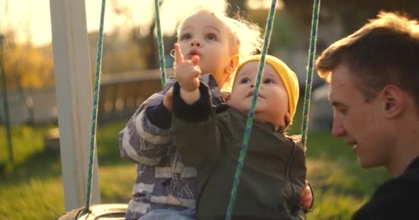慢动作：快乐的年轻父亲把他的儿子们推在秋千上。父亲和两个儿子在自家的院子里玩耍，秋千挂在树上，阳光灿烂。两个兄弟和父亲挥棒. — 图库视频影像