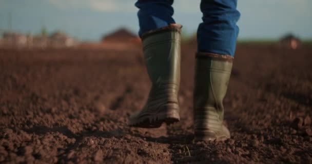 Un granjero cruza un campo con botas de goma sobre un fondo borroso del tractor en movimiento. Concepto de: Botas de goma, Estilo de vida, Agricultor, Moción lenta, Campos — Vídeo de stock