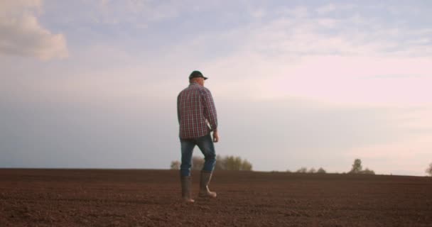 Un anziano agricoltore maschio con un cappello sta camminando attraverso un campo appena piantato. Guarda la terra fresca. Al rallentatore camminare a terra in stivali e guardando in lontananza . — Video Stock