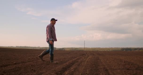 Un fermier âgé dans une casquette marche à travers un champ nouvellement planté. Regarde la terre fraîche. Au ralenti marcher sur le sol en bottes et en regardant dans la distance . — Video