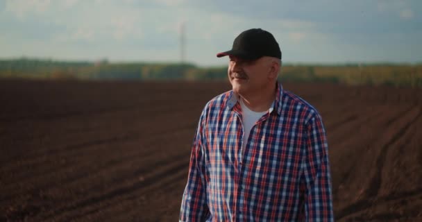A férfi farmer gumicsizmában megy a zöld mezőre a nap sugaraiban naplementekor. Agrártermesztés. Egy idős férfi farmer sétál egy újonnan ültetett mezőn. Nézd a friss földet. — Stock videók
