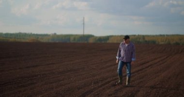 Erkek çiftçi gün batımında güneşin altında yeşil bir tarlada lastik botlarla dolaşır. Tarım ekimi. Yaşlı bir erkek çiftçi yeni ekilmiş bir tarlada yürüyor. Taze toprağa bak.