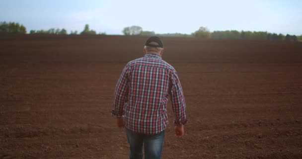 Un granjero anciano con gorra está caminando a través de un campo recién plantado. Mira la tierra fresca. En cámara lenta caminando en el suelo con botas y mirando a la distancia . — Vídeos de Stock